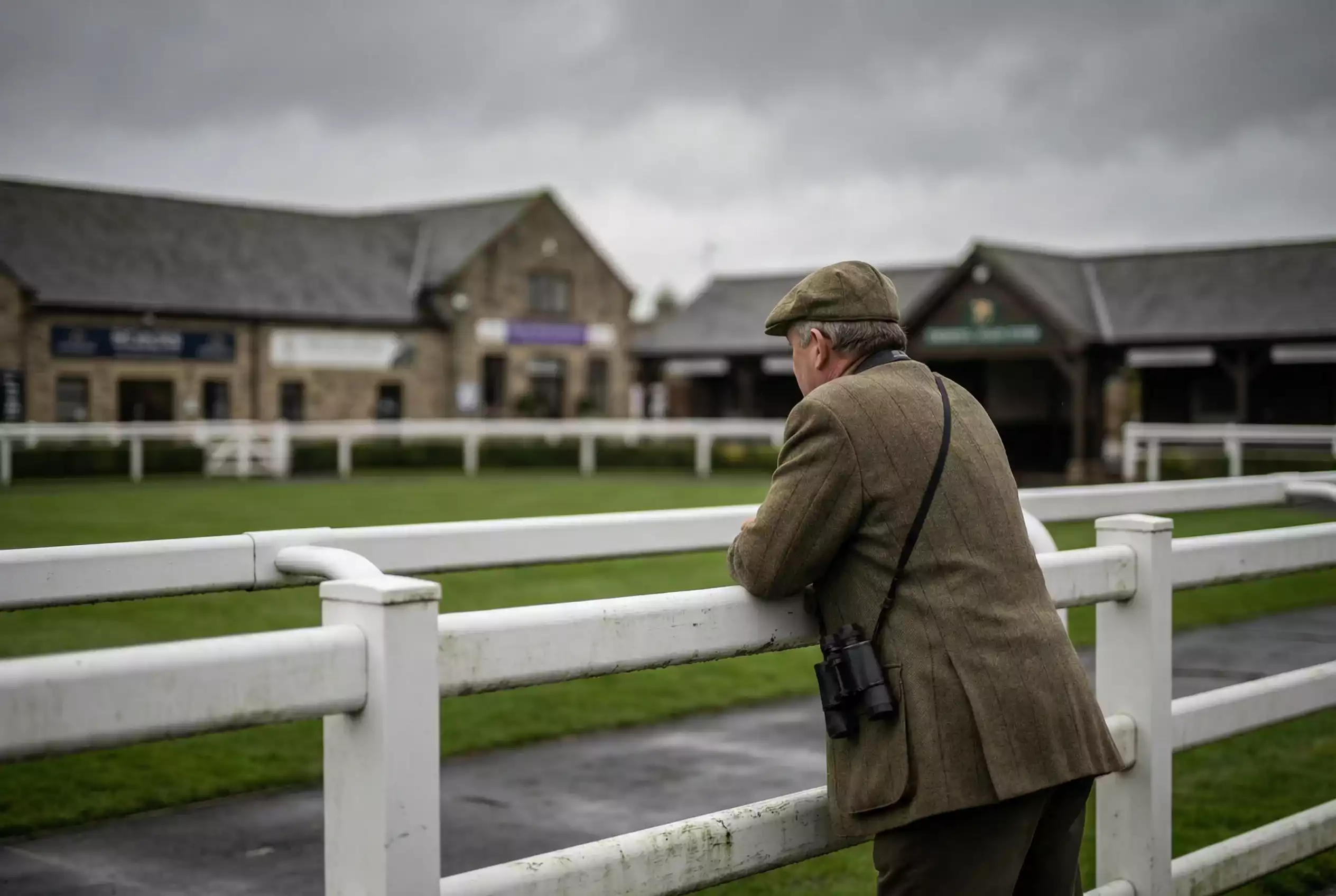Racehorse owner in elegant attire watching empty paddock with withdrawn horse